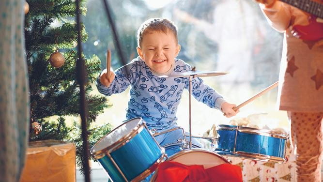 Ein ca. 5-jähriger Junge mit einem Grinsen auf dem Gesicht spielt am Schlagzeug mit einem Weihnachtsbaum im Hintergrund.