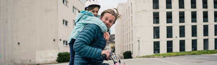 Ein Vater fährt mit dem Fahrrad seiner Tochter während dem sie mit dem Helm auf dem Kopf auf seinem Rücken sitzt.