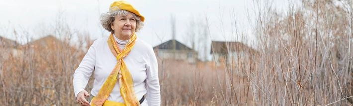 Eine ältere Frau in weissem Pullover und gelbem Schalt und Perret läuft durch ein Feld.