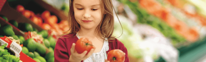 Ein kleines Mädchen ist im Supermarkt, hält in jeder Hand einen roten Apfel und sucht sich einen aus.