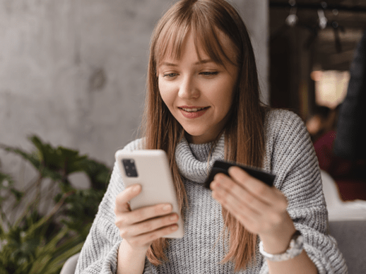 Eine junge Frau blickt erfreut auf ihr Smartphone und ihre Kreditkarte