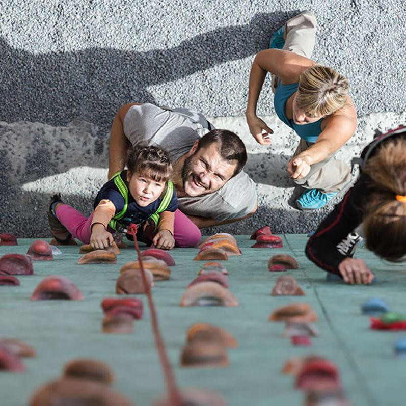 Immagine simbolica per la copertura dei rischi: una vista dall'alto verso il basso di un padre che sostiene la figlia attaccata alla corda mentre si arrampica su una parete in una palestra da arrampicata