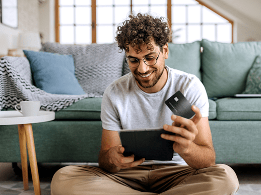 Un giovane ragazzo in salotto ha la sua carta di credito Platinum in mano mentre fa shopping online dal suo tablet